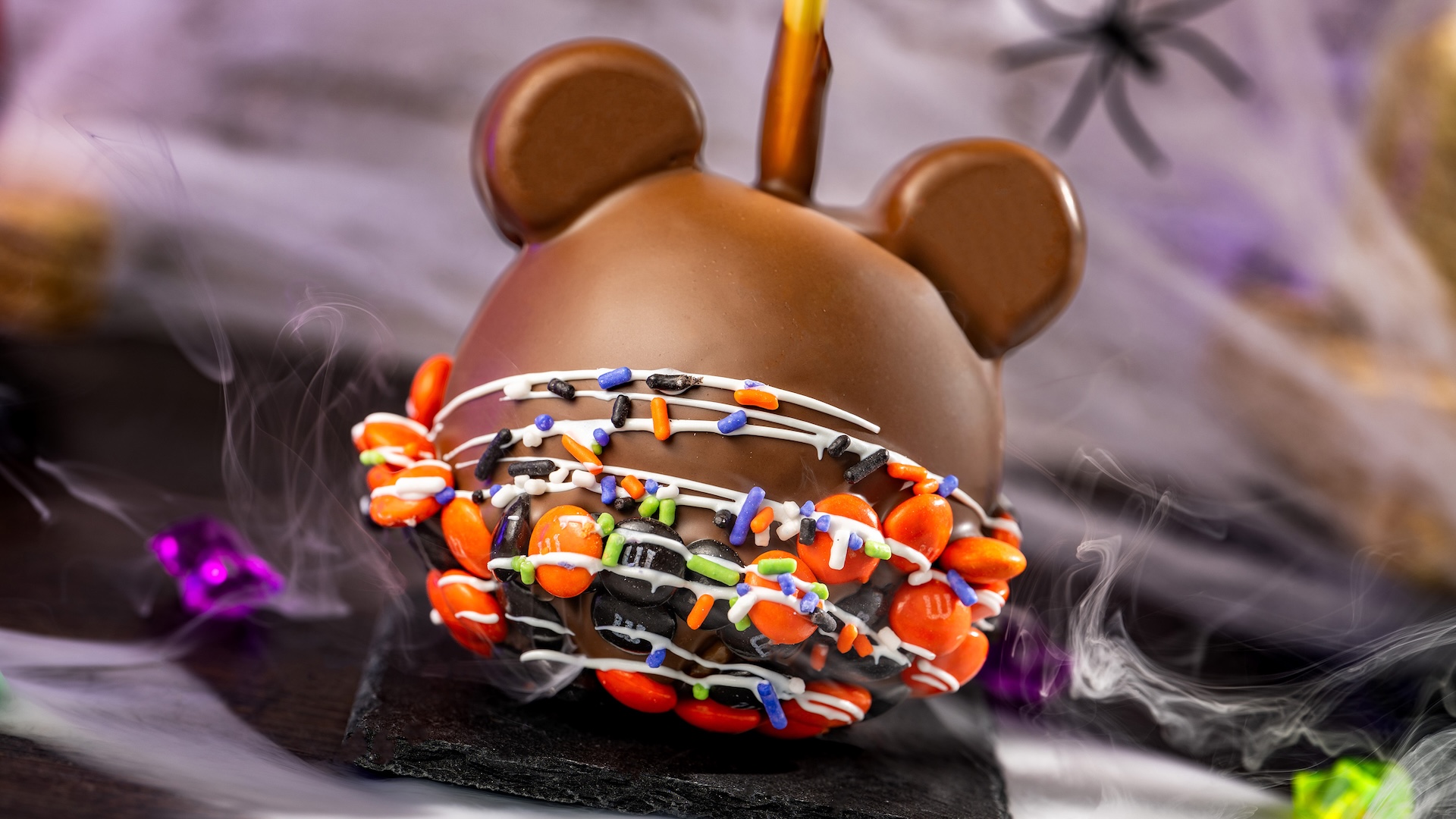 Mickey Halloween Caramel Apple with M&M’S Milk Chocolate Candies: Granny Smith apple with marshmallow ears dipped in caramel and milk chocolatey coating with M&M'S milk chocolate candies Halloween blend, Halloween sprinkles, and white chocolatey drizzle 