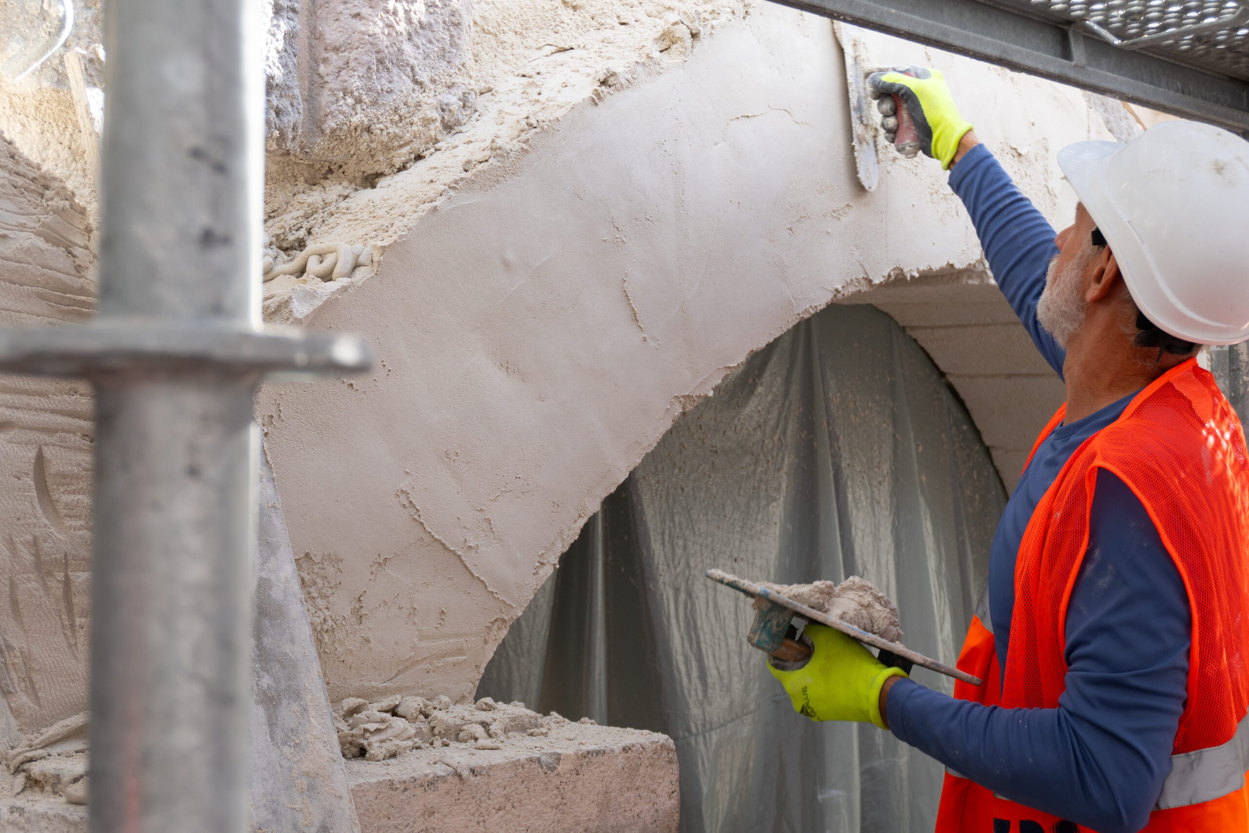 A worker in a hard hat smooths pale plaster onto an arch with a trowel, among scaffolding.
