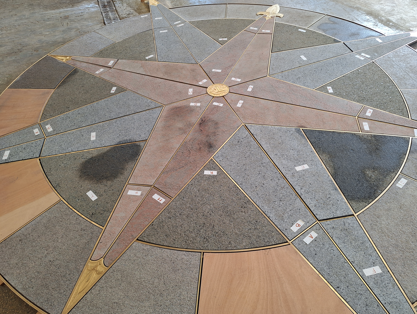 Granite compass rose with brass lines and gilded fleurs-de-lis, laid flat in a workshop with labelled pieces.